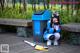 A woman sitting on a wooden bench next to a trash can.